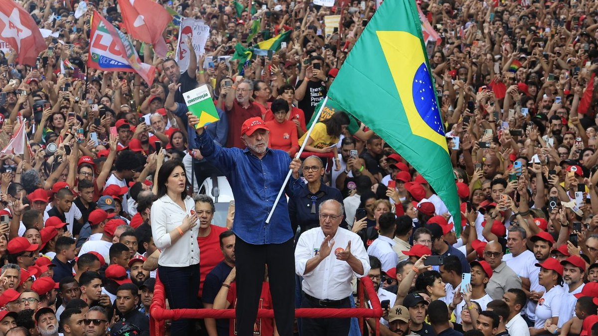 Lula, de camisa azul e boné vermelho, ergue a Constituição e uma bandeira do Brasil no encerramento da campanha do 2º turno, na Avenida Paulista, cercado por apoiadores.
